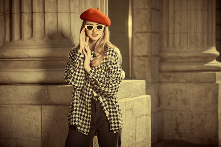 Portrait of a beautiful young woman with long wavy hair posing on a city street in plaid coat, red beret and beige sunglasses. Copy space. Elegant retro style in fashion.の写真素材
