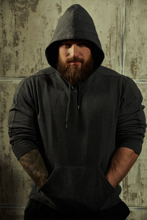 The concept of masculinity and physical strength. Portrait of a large brutal bearded man in a black hoodie looking at the camera. In the background is a concrete wall.の写真素材