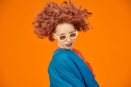 Positive Fashion. Portrait of attractive young woman fashion model with flying curly red hair posing emotionally in blue jacket and stylish beige sunglasses on a studio orange background. Copy space.の写真素材