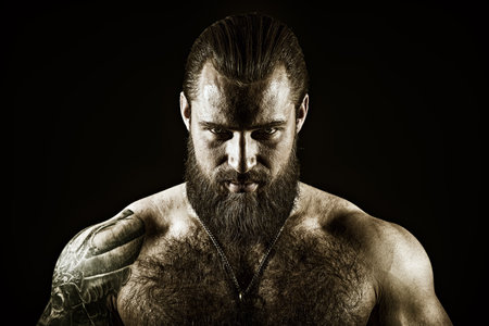 A brutal muscular tattooed bearded man with a naked torso looks confidently into the camera. Black studio background. Courage and physical strength. Bodybuilder portrait.の写真素材