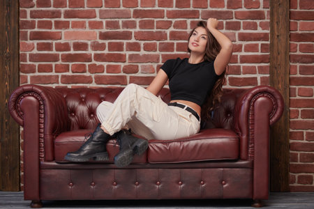Attractive confident young woman with beautiful long curly hair posing on a leather sofa in a black top, sporty white trousers and black boots. Beauty and fashion. Confident lifestyle.の写真素材