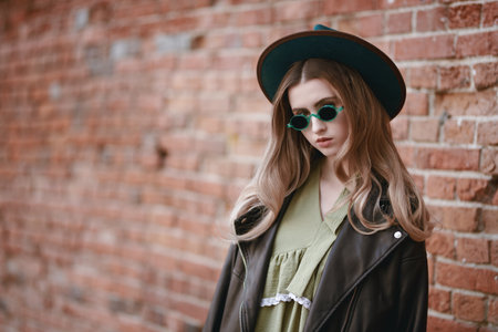 Beautiful hipster girl in stylish sunglasses, hat and leather jacket stands on a city street on brick wall background. Boho and hipster style. People and lifestyle. Copy space.の写真素材