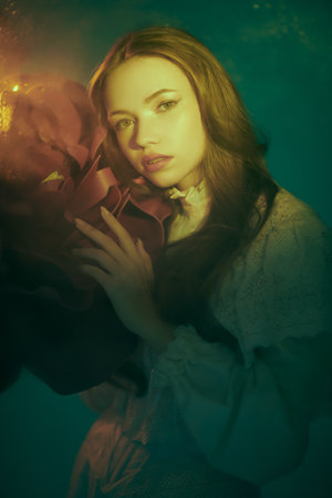 Art portrait of a beautiful tender girl with long hair, who stands in a warm light with a large peony flower in her hands. Fairy elf girl. Beauty and Fantasy.の写真素材