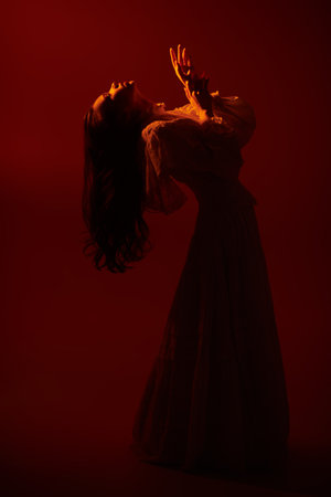 Shot of a beautiful romantic girl with long wavy hair posing in a white vintage dress. Dark red studio background.の写真素材
