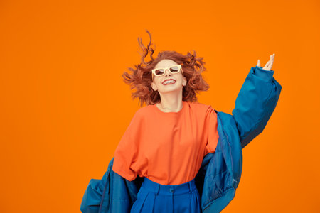 Portrait of beautiful cheerful girl with curly ginger hair smiling laughing in bright blue and orange clothes and beige sunglasses. Bright colors. Studio orange background. Place for text.の写真素材