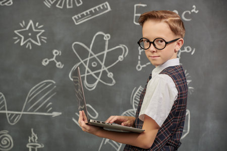 Shot of a smart boy in a school uniform and glasses looking at the camera seriously with a laptop. The background with a black chalkboard with scientific drawings. Education. Young scientist.の写真素材