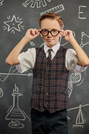 Portrait of a smart boy in a school uniform and glasses looking at the camera seriously. The background with a black chalkboard with scientific drawings. Education. Young scientists.の写真素材