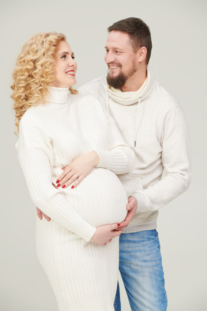 A loving husband and a beautiful pregnant wife are expecting a baby. Loving spouses in casual clothes hug their tummy with a baby on a white studio background.の写真素材