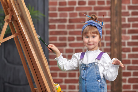 Art studio for children. Portrait of a happy little cute girl who paints on an easel with a palette and brushes in her hands and smiles. Creative development of children. Happy childhood.の写真素材