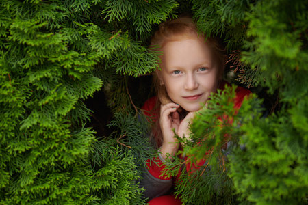 A cute little girl with red hair in a stylish jacket stands calmly among green bushes. Children's fashion.の写真素材