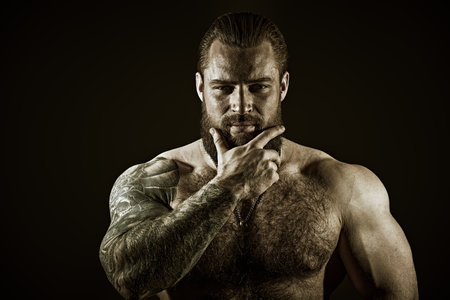 A brutal muscular tattooed bearded man with a naked torso looks confidently into the camera. Black studio background with copy space. Courage and physical strength. Bodybuilder portrait.の写真素材