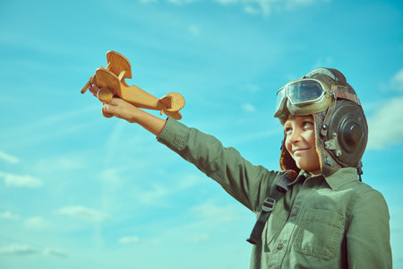A boy in aviator's helmet plays with a toy plane outdoors on a sunny day on the background of a blue sky. Children's games and dreams.の写真素材