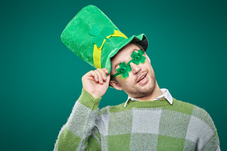 St. Patrick's day celebration. A handsome mature man wearing a  leprechaun hat and glasses in shape of clover leaves. Studio portrait on a green background, copy space.の写真素材