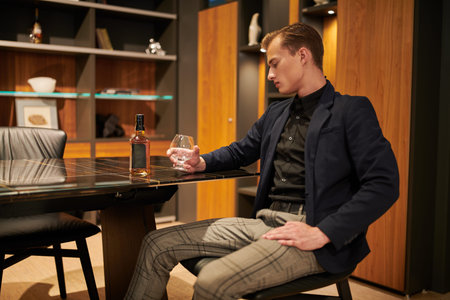 A thoughtful young businessman sits at the table in his respectable office drinking whiskey.