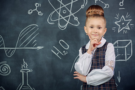 Portrait of a cute smart girl in a school uniform and glasses looking seriously at the camera. The background with a black chalkboard with scientific drawings. Young scientists. Educational concept.の写真素材
