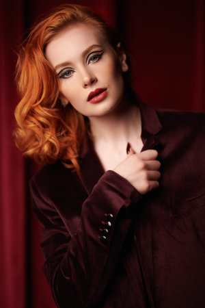 Elegant hairstyle. A charming red-haired young woman in an expensive velvet men's jacket in a dimly lit room. Fashion and femininity. Women's beauty and style. Elegant makeup with arrows.の写真素材