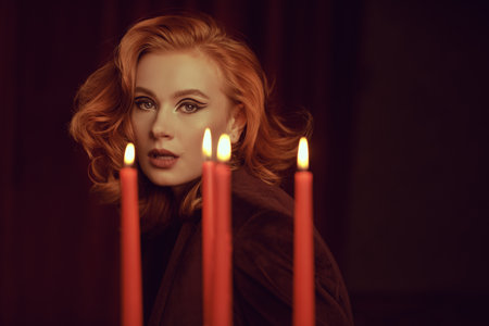 A charming red-haired young woman in an expensive velvet jacket against the background of red velvet curtains behind candles. Elegant hairstyle and makeup with arrows. Women's beauty and Fashion.の写真素材