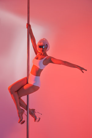 Dance on pole. Futuristic style. Cyber punk girl. Beautiful slender sexy girl dancer posing in white shorts, top, white wig and mirror glasses in a white interior in red light on pole.の写真素材