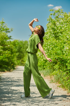 Summer fashion. Beautiful Asian girl posing in a trendy green overall and lemon sunglasses in a summer park with lush greenery around. Sunny day. Full length portrait.の写真素材