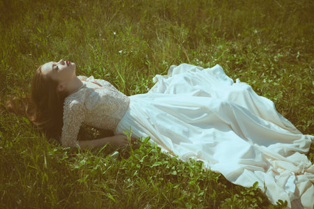 A gorgeous girl in a beautiful lace dress rests on the green grass on a sunny day. Summer freedom. Beauty and fashion.の写真素材