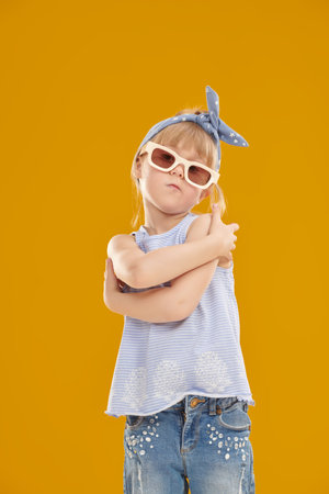 A cute five-year-old girl in summer clothes and fashionable sunglasses looks at the camera. Children's fashion. Yellow studio background with space for text. Children, happy childhood.の写真素材