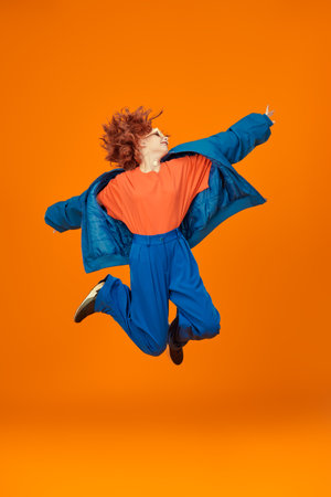Full-length portrait of attractive joyful fashion model girl with bright red curly hair jumping in blue and orange clothes. Bright colors. Studio orange background. Summer fashion.の写真素材