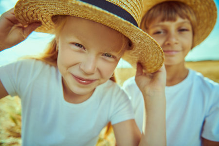 Two cute kids in white t-shirts and summer straw hats are in a filed on the background of a sunny blue sky. Summer time. Happy carefree childhood.の写真素材