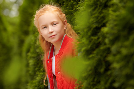 A red-haired little girl in a stylish jacket peeks joyfully out from behind the green bushes and smiles. Happy children. Kid's fashion.の写真素材