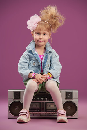 Pretty little girl singer in 80s style sits on a boombox and smiles. The child a pop and rock...
