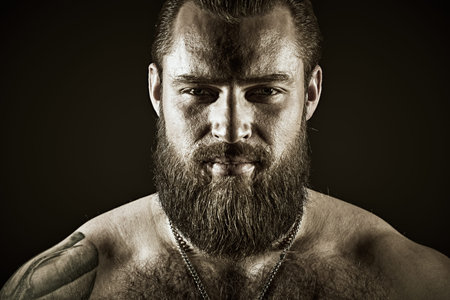 Bodybuilder portrait. A brutal muscular tattooed bearded man with a naked torso looks confidently...
