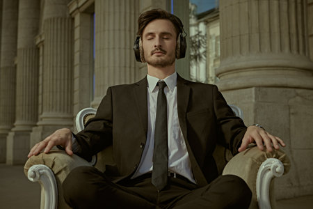 The concept of calmness and confidence. A young man in a business suit sits in an armchair on a city street with his eyes closed, wearing headphones on his head and with a slight smile.の写真素材