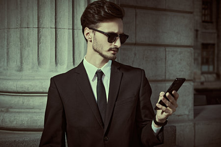 Business and communications. Portrait of a well-groomed young man in a classic black suit and black sunglasses, who is standing on a city street and using a smartphone.の写真素材