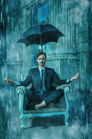 A well-groomed young man in a black suit with a white shirt and tie sits in an expensive chair on a city street with rainstorm under a black umbrella over his head. Business concept. Security and confidence.の写真素材