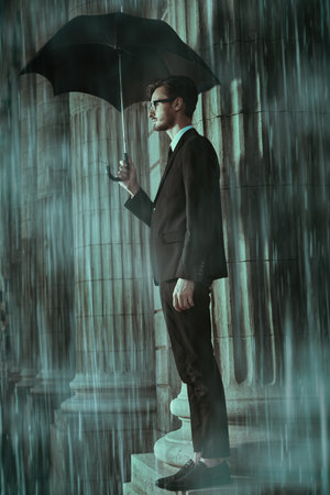 A handsome young man in a classic black suit with a white shirt, a tie stands in heavy rain under an umbrella at the columns on a city street. Businessman.の写真素材