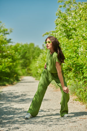 Summer fashion. Beautiful Asian girl posing in a trendy green overall and lemon sunglasses in a summer park with lush greenery around. Sunny day. Full length portrait.の写真素材
