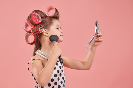 Kid's Fashion - clothes and accessories. A little pretty girl in an elegant polka-dot dress and curlers on her head is going to a party and doing her make-up. Pink studio background. Pin-up style.の写真素材