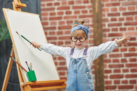 Portrait of a little cute girl in glasses who paints on an easel with brush in her hands. Creative development of children. Happy childhood. Art studio for children.の写真素材