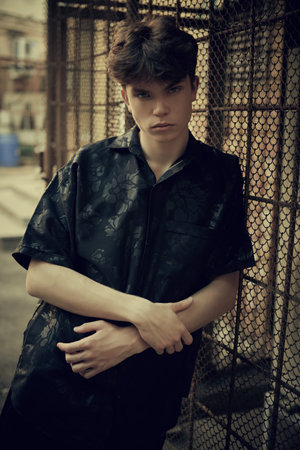 A modern young man in black clothes stands leaning against a metal grid in a dark alley of a big city. Urban lifestyle.の写真素材