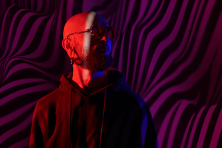 Expression of personality. Portrait in mixed color light of a bald, bearded brutal man, wearing a black hoodie and round glasses. Studio shot.の写真素材