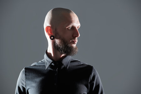 An informal shaven-headed bearded man with a brutal appearance poses against a grey studio background. Copy space.の写真素材