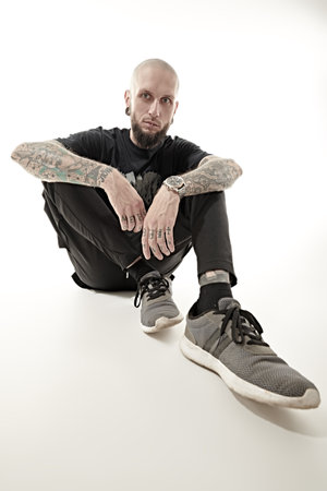 Rock musician. A man with an informal appearance - a shaved head and with piercings on his lips and ears, with a tattoo on his body - poses at the studio in black clothes. White studio background.の写真素材