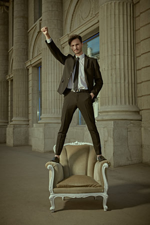Success. A successful and confident young man in a business suit stands on an armchair on a city...