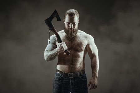 A handsome muscular tattooed man with a beard stands with a large ax in his hand and poses confidently. Dark gray studio background. Courage and physical strength.の写真素材