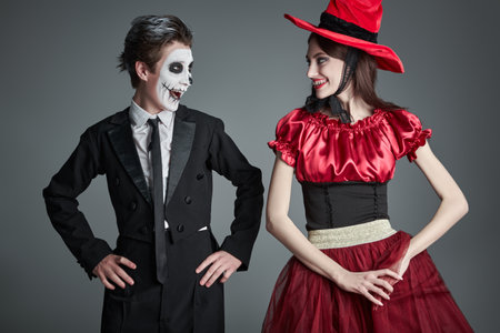 A teenage boy in a costume of sugar skull boy and a girl in a witch's costume posing together with fun at studio. Grey background. Halloween party for teenagers.の写真素材