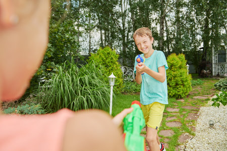 Two joyful kids playing with water guns in the garden on a summer day. Summer joy and children's games.の写真素材