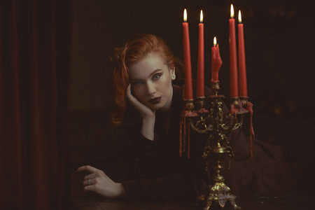 Charming red-haired young lady in a burgundy velvet jacket stands with candles in a dark room. Elegant hairstyle and makeup with arrows. Women's beauty and Fashion.の写真素材