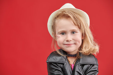 Portrait of a little cute girl with glitter makeup, in fashionable clothes and accessories of a pop and rock musician on red studio background with copy space. Children's fashion. Little fashion-girl.の写真素材