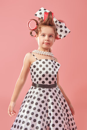 Pin-up style. A little pretty girl in an elegant polka-dot dress and curlers on her head is going to a party. Pink studio background. Kid's Fashion - clothes and accessories.の写真素材
