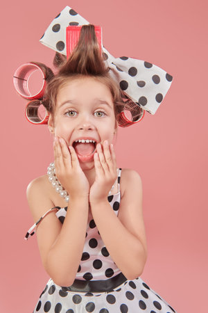 An emotional little pretty girl in an elegant polka-dot dress and curlers on her head screams in surprise. Pink studio background. Kid's Fashion - clothes and accessories. Pin-up style.の写真素材