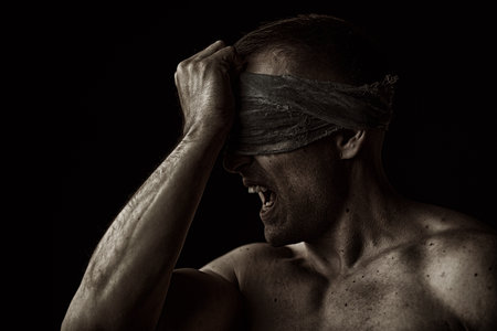 Psychological picture. A large, strong man with a naked torso stands with a gauze bandage over his eyes and screams. Studio portrait on a black background with copy-space.の写真素材